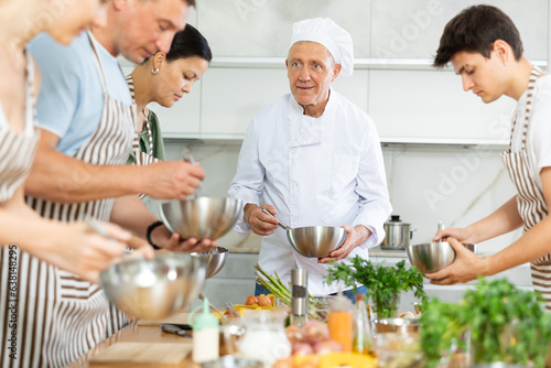 Smiling elderly man  seasoned professional chef conducting culinary courses  imparting cooking skills to diverse group of people of different ages