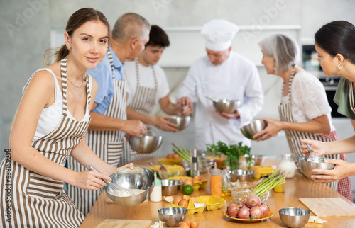 Positive interested young woman attending group culinary classes  standing by table with ingredients and utensils  learning culinary skills from professional chef