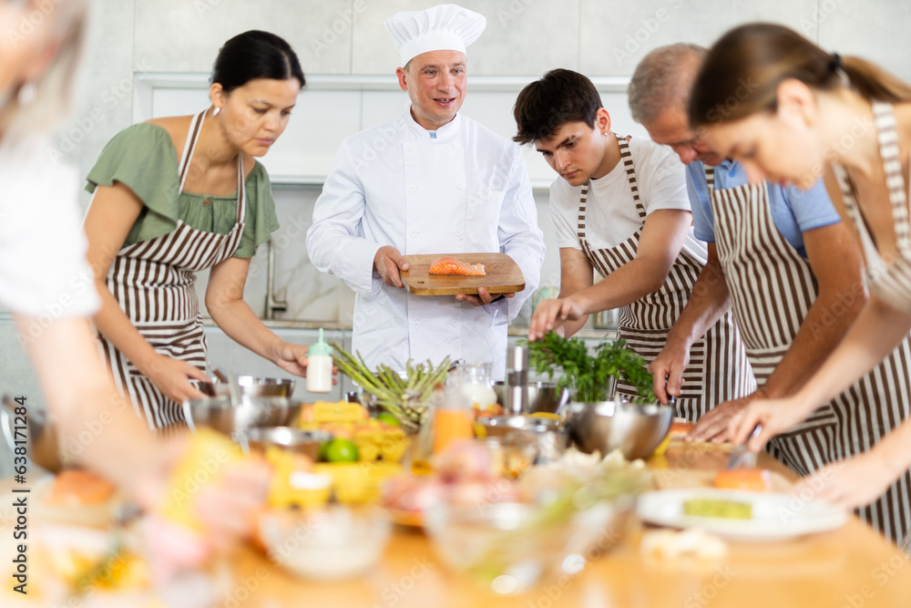 Friendly experienced male chef in white uniform giving culinary classes to group of men and women of different ages, teaching to cook salmon deliciously