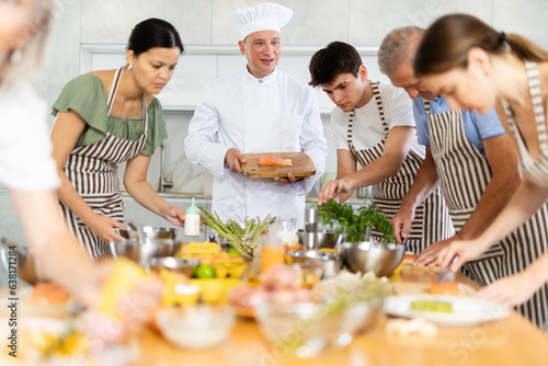 Friendly experienced male chef in white uniform giving culinary classes to group of men and women of different ages  teaching to cook salmon deliciously
