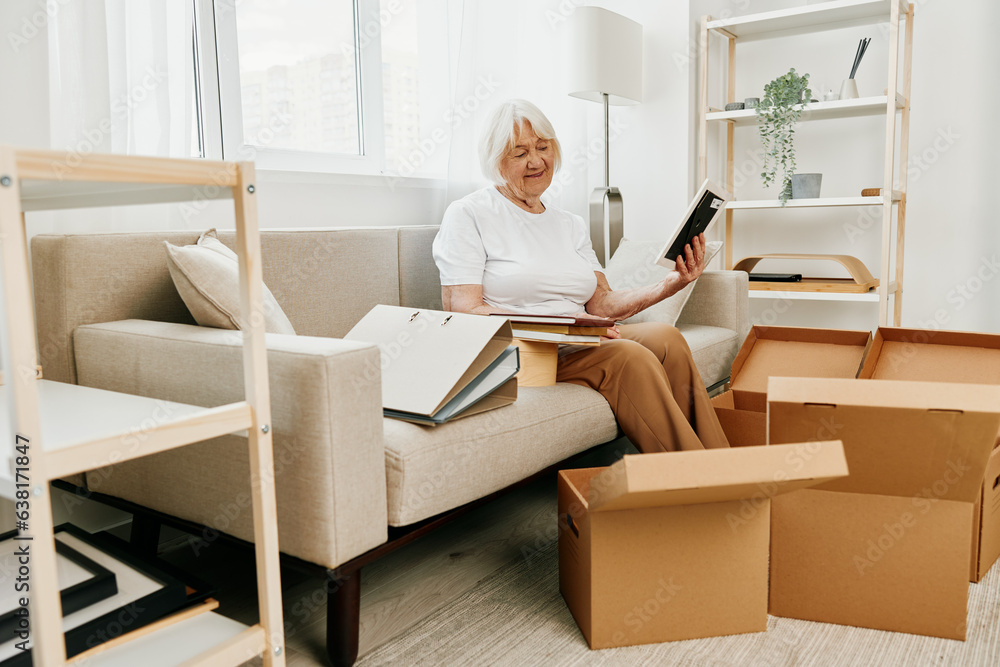 elderly woman sits on a sofa at home with boxes. collecting things with memories albums with photos and photo frames moving to a new place cleaning things and a happy smile. elderly woman sits on a sofa at home with boxes. collecting things with memories albums with photos and photo frames moving to a new place cleaning things and a happy smile.