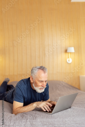 Handsome confident senior bearded man freelancer lying on bed using laptop in living room