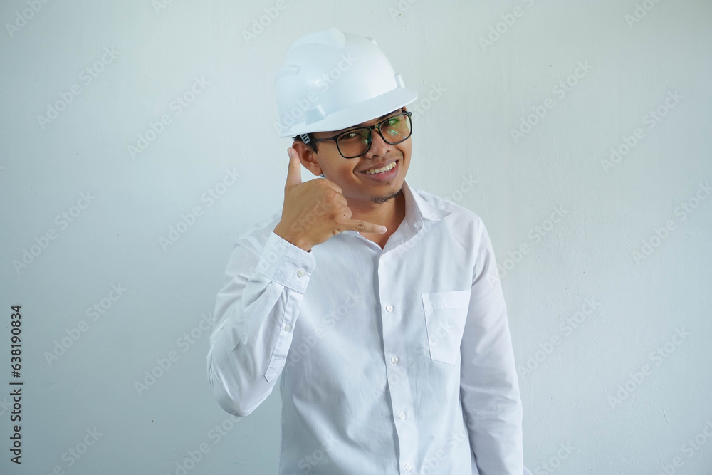 young asian man is engineer wearing helmet standing with showing a mobile phone call gesture with fingers and smiling with confident, architect or contractor, worker or labor, industrial concept.