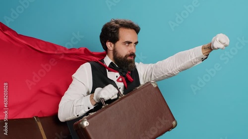 Hotel bellhop acting as superhuman with cape, offering to help people with suitcase luggage in studio. Young adult doorman presenting comic hero character with red fabric, feeling determined. photo