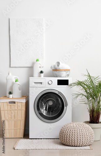 Washing machine with bottles of detergent  laundry basket and pouf near white wall