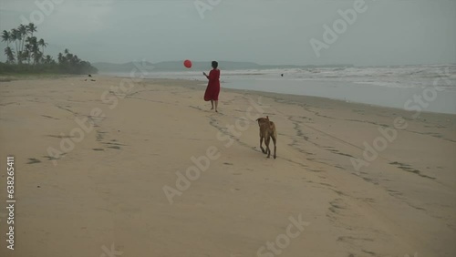 Cinematic shot of an indian fashian model running on a sandy beach with a red dress and a red balloon in her hand while a dog is walking behind her in Goa India photo