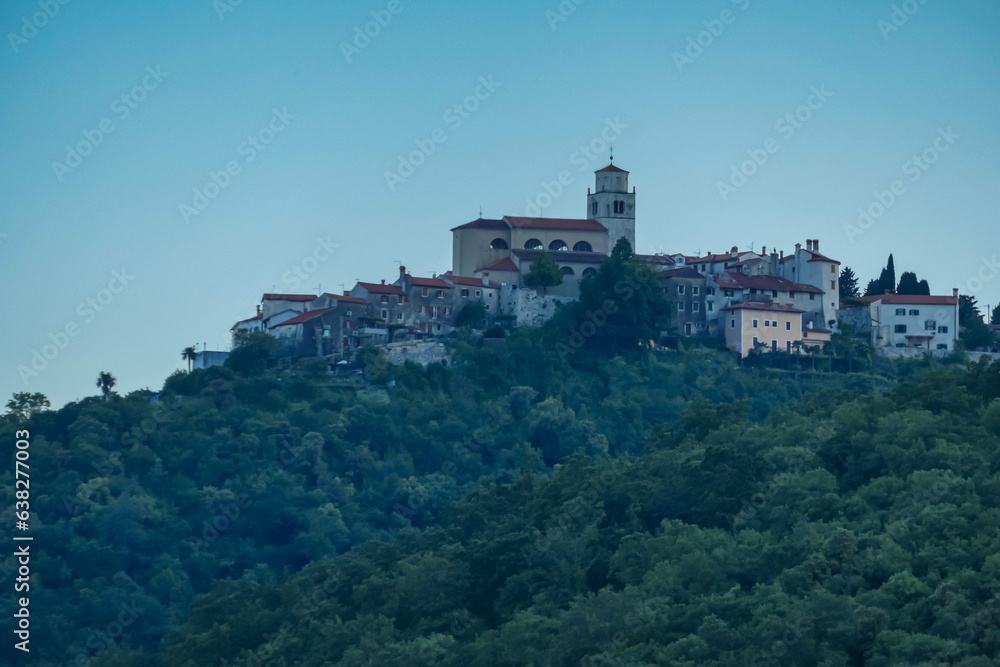 A distant view on a small town located at the top of the hill in Moscenicka Draga, Croatia. A tall church tower is towering above the other buildings. Lush forest overgrowing the hills. Dusk.