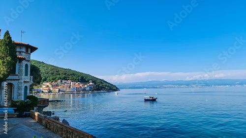 A panoramic view of the shore along Moscenicka Draga, Croatia. There is a small town located on the shore of the Mediterranean Sea. Few boats crossing the calm sea. Green hills in the back. Sunny day.