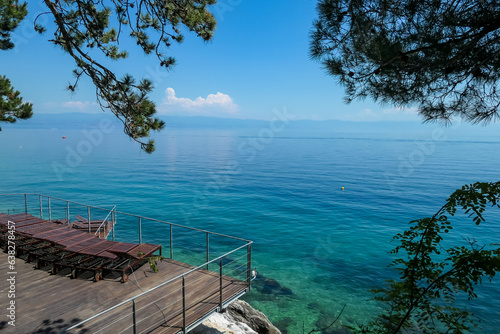 A small resting area at the shore of Mediterranean Sea in Moscenicka Draga, Croatia. There are a few tree branches in the frame. Mountain chains visible in the back.  Calm sea. Clear and sunny day. photo