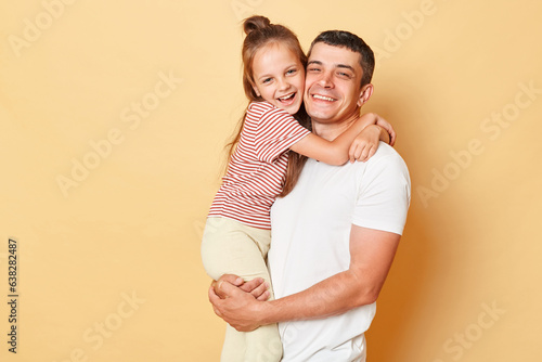 Smiling brunette man holding his little daughter, child hugging her father family looking at camera with happy faces standing in casual clothing isolated over beige background.