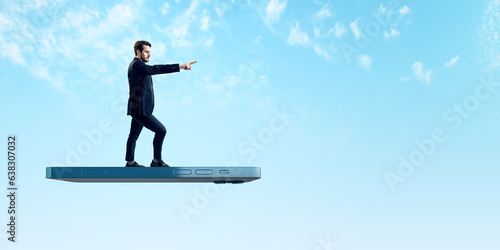 User experiences for mobile concepts. Young businessman pointing forward on huge flying smartphone on wide bright blue sky background with mock up place.
