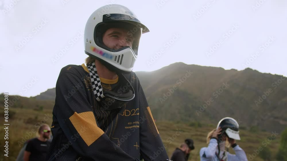 Amidst a stunning mountainous landscape, a young man sits astride a vintage off-road motorcycle, donning a white helmet that reveals his adventurous spirit. The open-faced helmet allows the wind to Amidst a stunning mountainous landscape, a young man sits astride a vintage off-road motorcycle, donning a white helmet that reveals his adventurous spirit. The open-faced helmet allows the wind to
