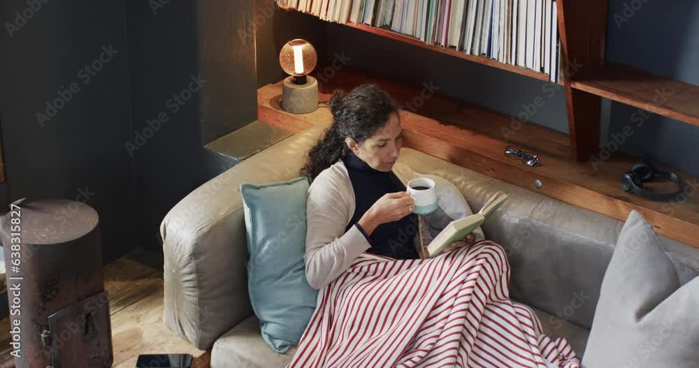 Biracial woman lying on sofa under blanket reading book and drinking coffee at home, slow motion Biracial woman lying on sofa under blanket reading book and drinking coffee at home, slow motion