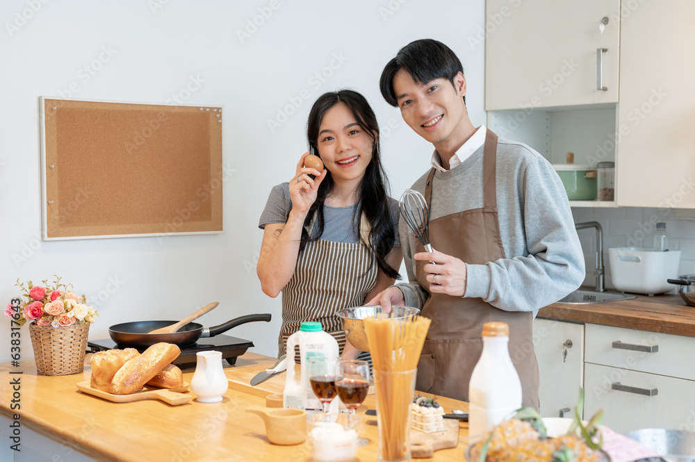 Fototapeta premium A lovely Asian couple in an apron stands in their kitchen together, cooking or baking pastry