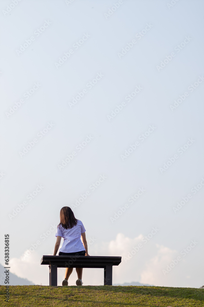 Back view of young woman sitting on long bench alone in evening looking at beautiful summer landscape background of nature and with copy space for text. concept loneliness of woman who is alone