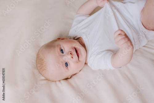 Portrait of happy funny cute little newborn boy with smiling face lying on comfortable bed in living room at home. Cheerful pretty infant baby resting playing lying down on blanket. Childhood concept