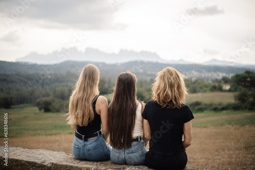 Three girls on their backs of different nationalities and ethnicities, sitting looking at the landscape, it is a field with the gray sky