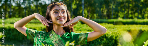 smiling young indian woman with bindi and sari posing near plants in park in summer, banner photo