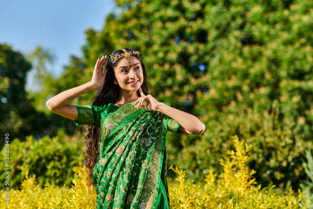 trendy young indian woman in traditional outfit and bindi posing near plants in park in summer trendy young indian woman in traditional outfit and bindi posing near plants in park in summer