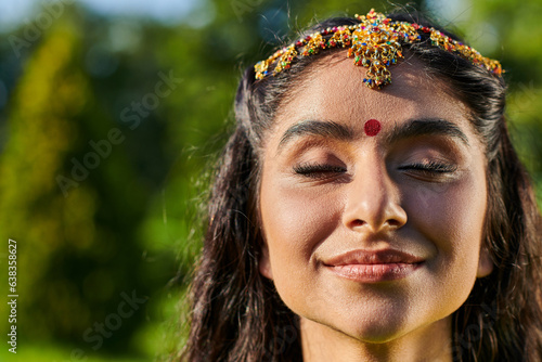 portrait of smiling indian woman with bindi and matha patti standing with closed eyes outdoors photo