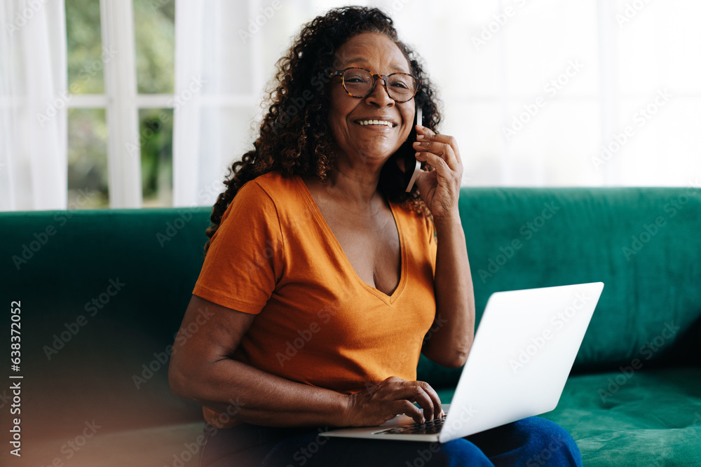 Fototapeta premium Mature woman making a phone call as she works from home with a laptop
