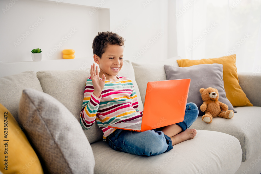 Photo of fun primary school kid sitting couch have online web lesson greetings classmates with netbook room indoors Photo of fun primary school kid sitting couch have online web lesson greetings classmates with netbook room indoors