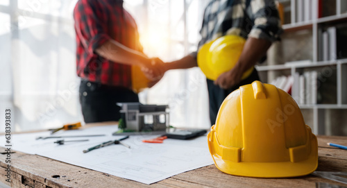 Yellow safety helmet on workplace desk with construction worker team hands shaking greeting start up plan new project contract in office at construction site, partnership and contractor concept photo