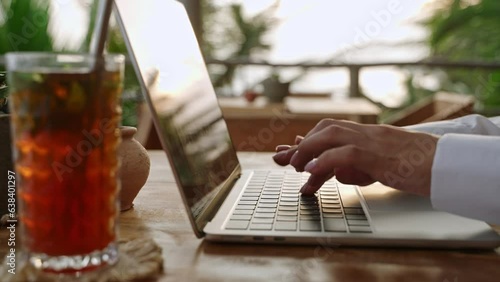 Woman typing on laptop keyboard in cafe. Writer creates a poem. Person composing email using notebook in outdoor coworking with ice tea, palm trees. Journalist writing article. Blogger making smm post