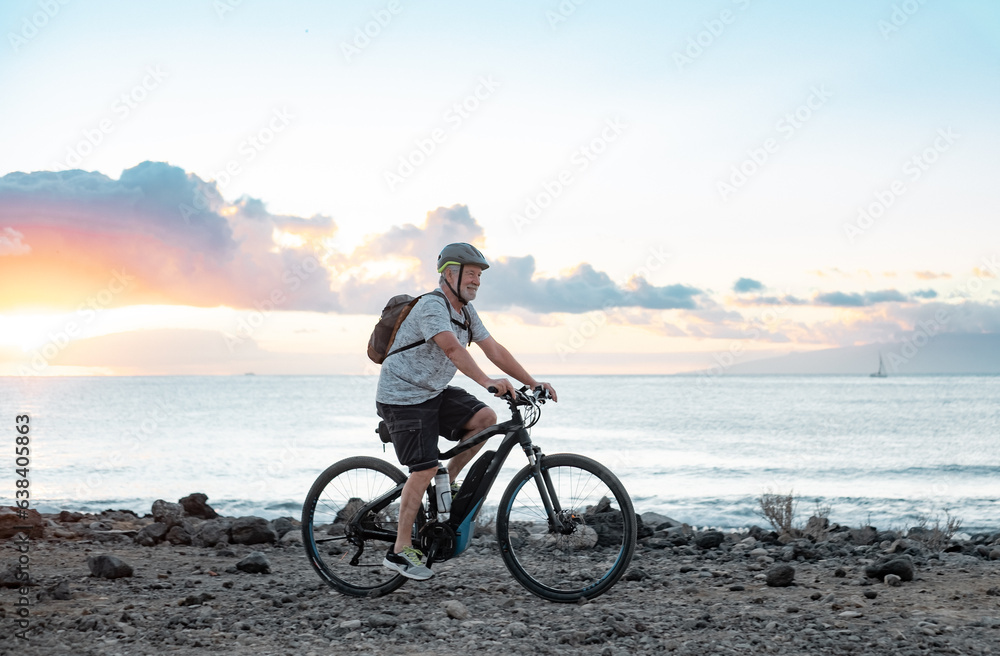 Fototapeta premium Happy active senior cyclist man at sea at sunset light with electric bicycle running on the beach - elderly man with helmet enjoying healthy lifestyle and freedom
