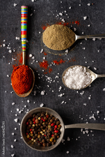 Old spoons with peppercorn, ground pepper, salt and red paprika powder on black background