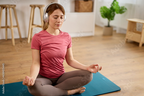 Beautiful young woman with headphones meditating on yoga mat at home