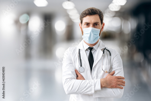 Photo of a caucasian proud male doctor, therapist, surgeon, cardiologist in a medical uniform, protective mask and with stethoscope on his shoulders, standing in a hospital, looking to the camera