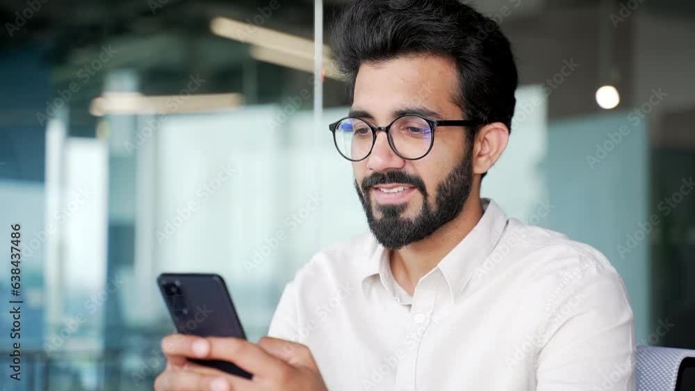 Close up. A smiling young businessman in a white shirt is using, browsing smartphone while sitting in modern office. Bearded entrepreneur in glasses checking email or writing text message to client