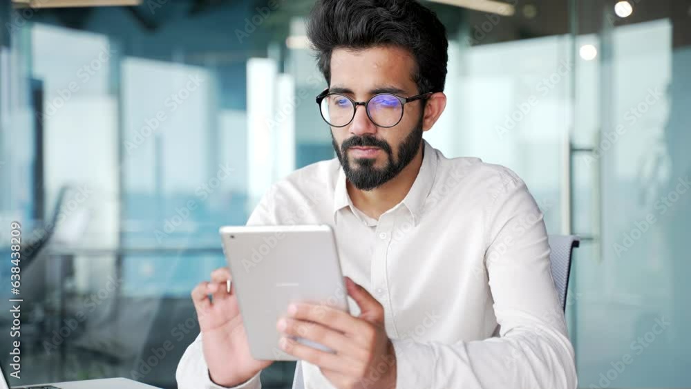 Close up. Serious young businessman in a white shirt is using, browsing a tablet while sitting in office. Entrepreneur in glasses looks at information on the Internet or writing text message to client Close up. Serious young businessman in a white shirt is using, browsing a tablet while sitting in office. Entrepreneur in glasses looks at information on the Internet or writing text message to client