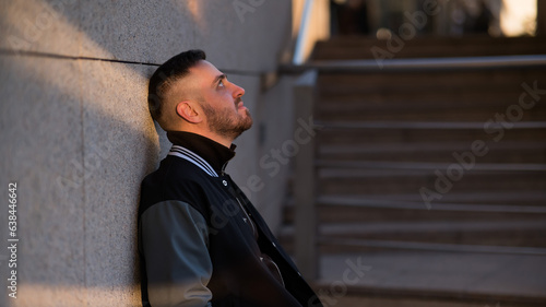 Caucasian bearded man in a bomber jacket leaned against a wall. 