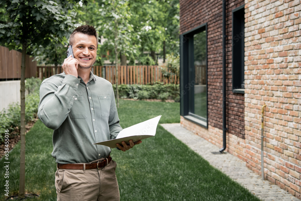Fototapeta premium cheerful real estate broker with folder talking on smartphone and looking at camera near house