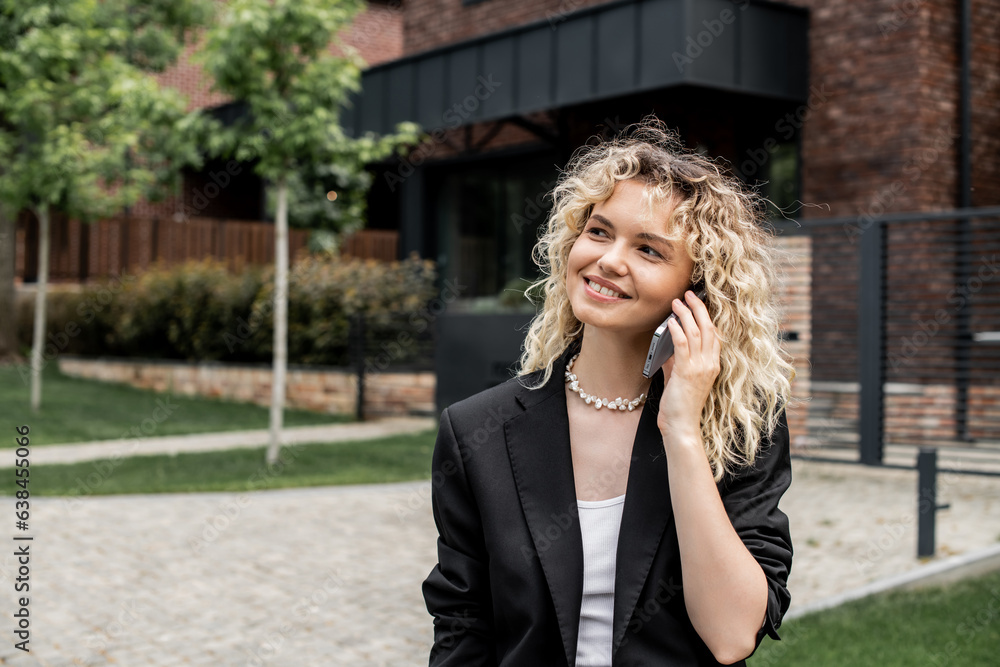 smiling property realtor looking away while talking on smartphone near building on city street