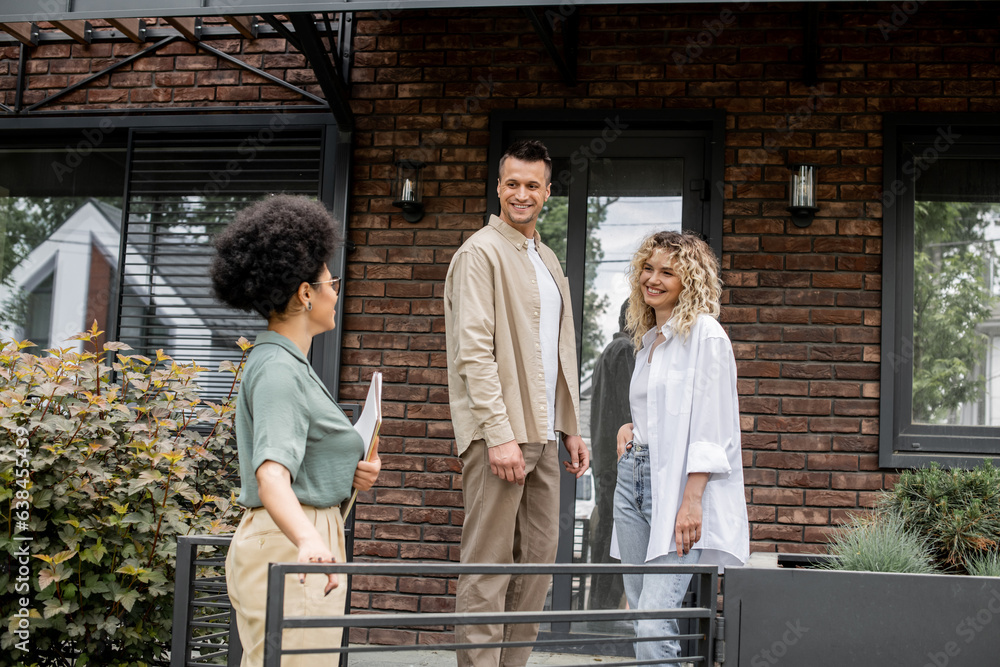 african american real estate agent talking to smiling couple near new modern cottage for sale