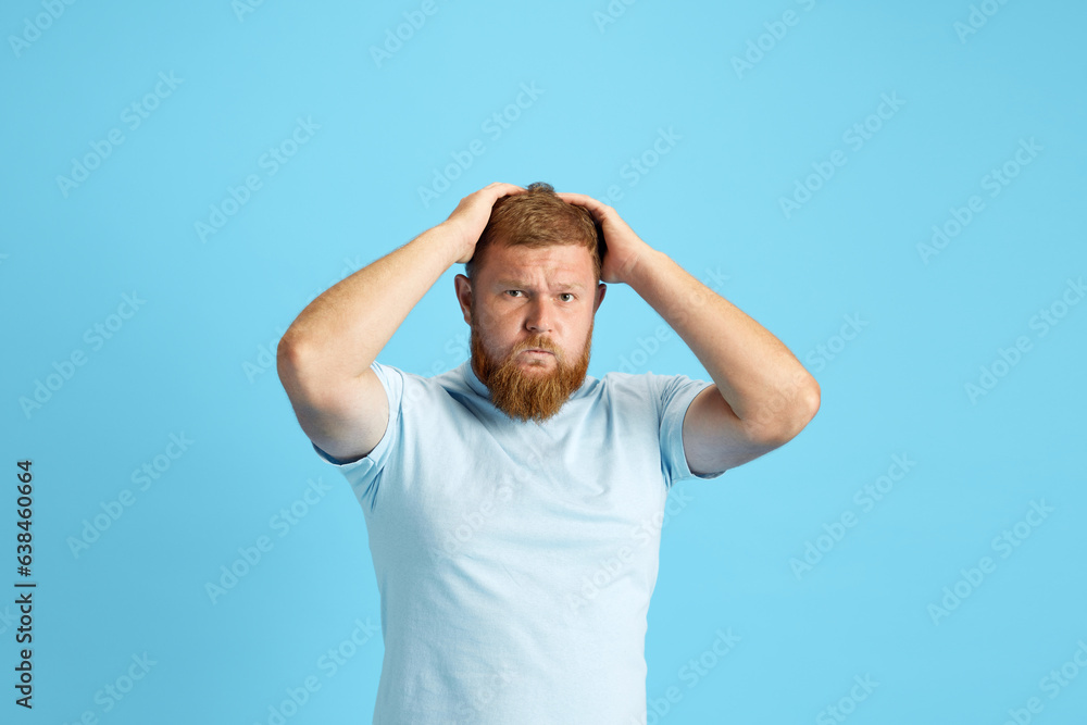 Emotional bearded man holding head with hands standing with shocked and stressful face against blue studio background. Concept of human emotions, lifestyle, facial expression, ad. Copy space for ad Emotional bearded man holding head with hands standing with shocked and stressful face against blue studio background. Concept of human emotions, lifestyle, facial expression, ad. Copy space for ad