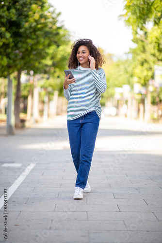 Full body happy young woman walking with mobile phone and earphones