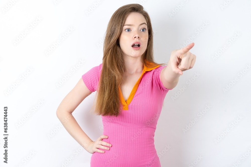 young beautiful red haired woman wearing pink dress Pointing with finger surprised ahead, open mouth amazed expression, something on the front. young beautiful red haired woman wearing pink dress Pointing with finger surprised ahead, open mouth amazed expression, something on the front.