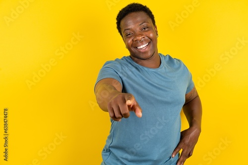 Young handsome man standing over yellow studio background pointing at camera with a satisfied, confident, friendly smile, choosing you photo