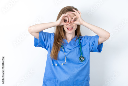 Beautiful doctor woman wearing blue medical uniform over blue background doing ok gesture like binoculars sticking tongue out, eyes looking through fingers. Crazy expression. photo