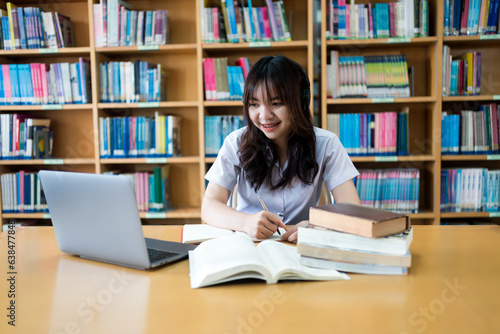 Young Asian female university student concentrate on studying, doing assuagement project in library with books and laptop on table