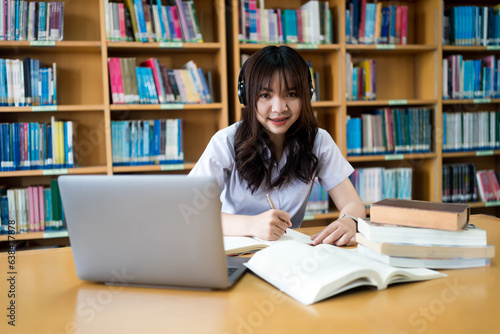 Young Asian female university student concentrate on studying, doing assuagement project in library with books and laptop on table
