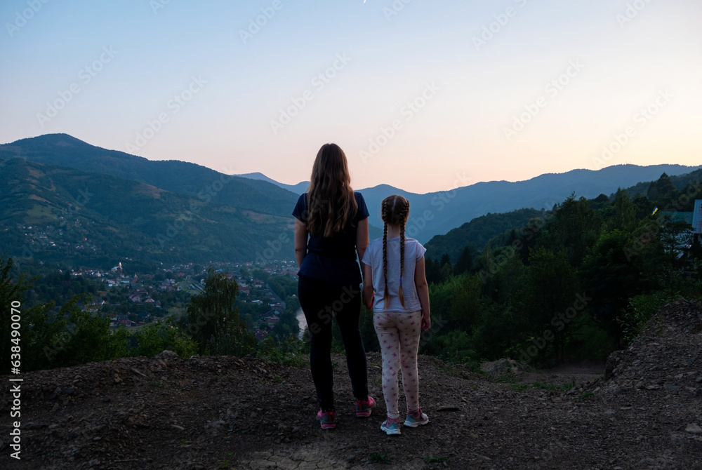 Travelers mother and daughter standing back view beautiful Carpathian Mountains background on sunset, having fun spending time together enjoying nature. Active lifestyle family travel tourism concept.