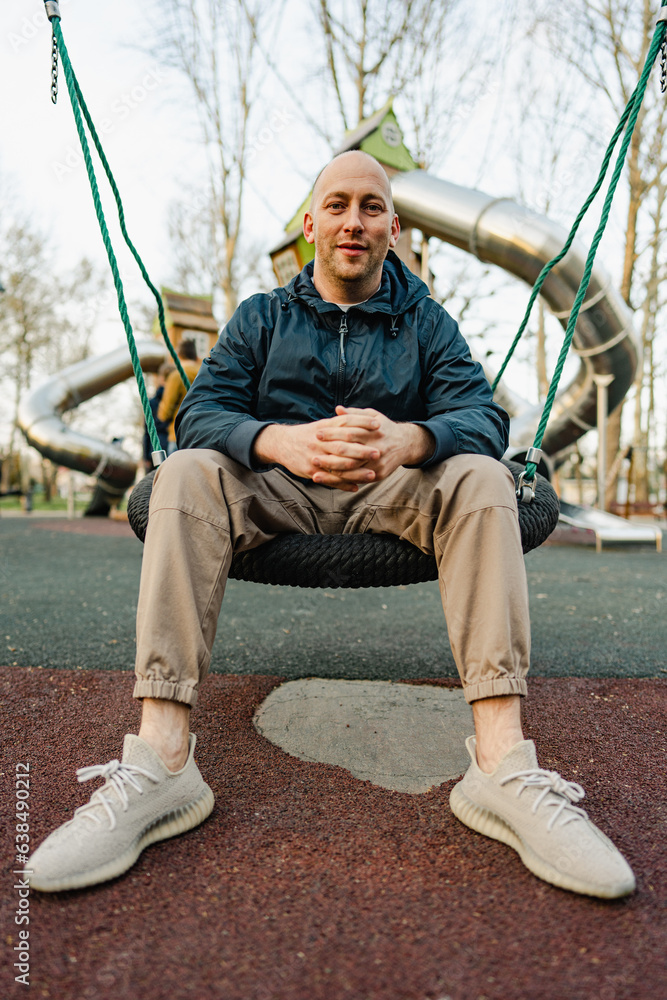 Smiling bald man sitting on a swing in a playground Smiling bald man sitting on a swing in a playground