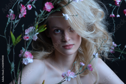 Beautiful woman with big wavy hair and purple make-up studio portrait. Model surrounded with flower twigs with blossoms with naked shoulders looking at camera. Toned image with blue color