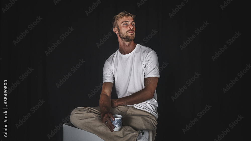 Young handsome blond man in casual t-shirt and beige pants on black background and sitting, posed seriously sipping a drink - obrazy, fototapety, plakaty 