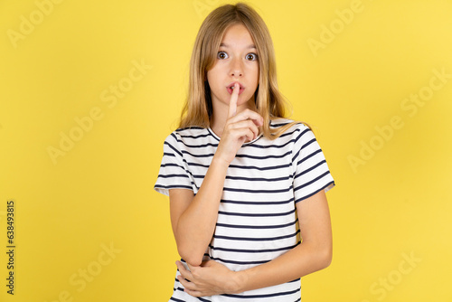 Surprised Beautiful caucasian kid girl wearing striped  T-shirt makes silence gesture, keeps finger over lips and looks mysterious at camera photo